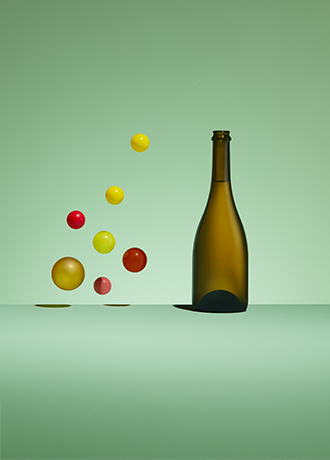 Bouteille champenoise en verre vert antique, modèle Millésime, à côté de sphères colorées jaunes et rouges flottant dans l'air sur un fond vert pastel.