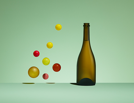 Bouteille champenoise en verre vert antique, modèle Millésime, à côté de sphères colorées jaunes et rouges flottant dans l'air sur un fond vert pastel.