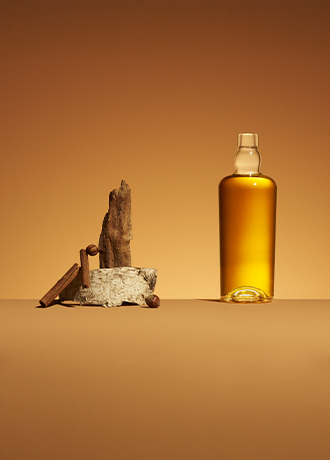 Bottle of amber spirit, Malty model, next to a composition of barks, cinnamon sticks, and hazelnuts on an ochre background.