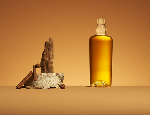 Botella de licor ámbar, modelo Malty, junto a una composición de cortezas, ramas de canela y avellanas sobre un fondo ocre.