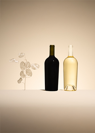Bouteille de vin rouge en verre vert et bouteille de vin blanc en verre blanc, modèle Antik Eco, à côté d'une branche de monnaie-du-pape sur un fond beige.
