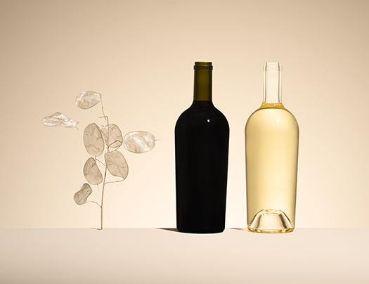 Bouteille de vin rouge en verre vert et bouteille de vin blanc en verre blanc, modèle Antik Eco, à côté d'une branche de monnaie-du-pape sur un fond beige.