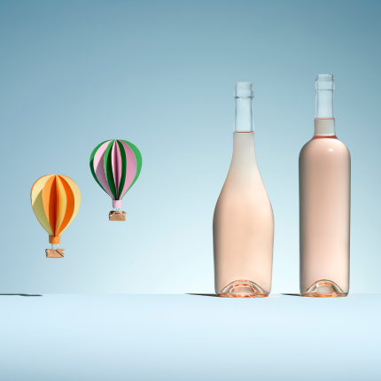 Dos botellas de vidrio blanco de diferentes formas para vino rosado, junto a dos coloridos globos aerostáticos de papel sobre un fondo azul pastel.