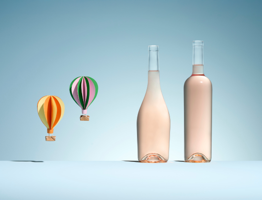 Deux bouteilles en verre blanc de formes différentes pour vin rosé, à côté de deux montgolfières colorées en papier sur un fond bleu pastel.